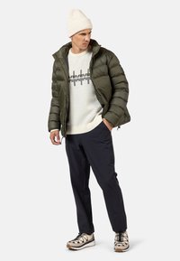 Olivgrüne Steppjacke mit einem Reißverschluss vorne, über einem cremefarbenen Pullover mit schwarzen Wellenmustern getragen. Kombiniert mit marineblauen Hosen und beigen Schuhen.