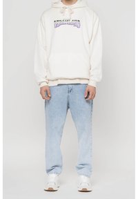 Krémová mikina s potiskem na přední straně, kapsou na přední straně a kapucí se stahovací šňůrkou; spárovaná se světle modrými volnými džíny a bílými teniskami s hnědými podrážkami.