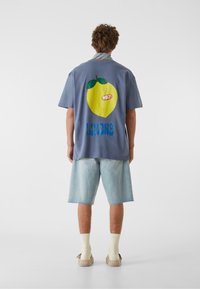 Lyseblå bomullsskjorte med korte ermer, som har et stort grafikk av en gul sitron og "LIMONE"-tekst på baksiden, kombinert med lyse denimshorts.