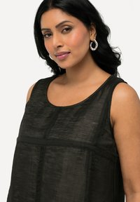 Top negro sin mangas con una tela texturizada, escote redondo y costuras verticales. La modelo lleva puestos pendientes grandes de aro plateados.