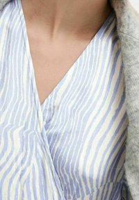 Modrý a bílý pruhovaný top s designem V výstřihu, vyrobený z texturované látky. Ukazuje jemný lesk a splývavost, vrstvený s svetle šedým kouskem oblečení.