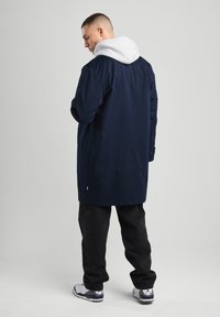 Navy blue long coat with a gray hood, featuring a straight cut and side pockets. Worn over black pants and white sneakers.