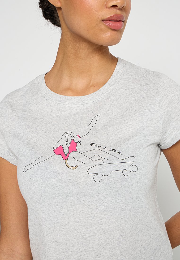 Frau trägt ein hellgraues Shirt mit einer Linzeichnung einer Eiskunstläuferin in einem pinken Outfit und dem Text "Feuer & Eis" auf der Brust.