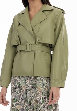 Femme portant une veste en cuir vert clair ceinturée avec de larges revers, sur une jupe à motif floral.