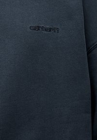 Σκούρο γκρι φούτερ από υφή υφάσματος με στρογγυλή λαιμόκοψη. Διαθέτει διακριτικό, ανάγλυφο λογότυπο "Carhartt" στο στήθος.