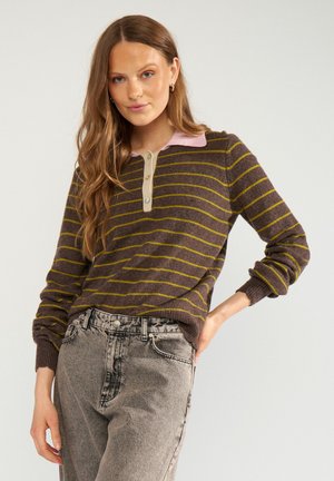 Femme aux longs cheveux portant un pull à rayures marron avec un col rose et un jean gris délavé taille haute, debout devant un fond uni.
