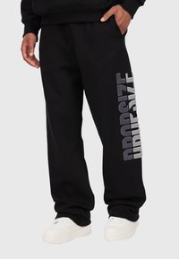 Schwarze Jogginghose aus weichem Material mit lockerer Passform. Das linke Bein zeigt "DROPSIZE" in fetten, silbernen Buchstaben.