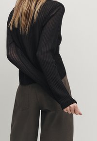 Schwarzes, geripptes Langarmoberteil mit taillierter Passform. Der Stoff wirkt leicht und leicht durchsichtig. Kombiniert mit hochgeschnittenen, olivgrünen Hosen.