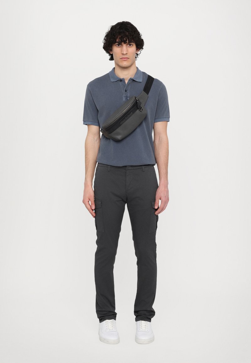 Uomo che indossa una polo grigia, pantaloni cargo neri e sneakers bianche. Porta una borsa a tracolla nera e dal design elegante. Design semplice e moderno.