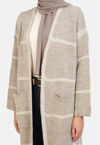 Cardigan en tricot beige avec un motif rayé blanc, à l'avant ouvert, avec deux poches avant et des manches longues, porté sur un haut clair et un jean.