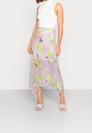 Kvinde iført hvid ærmeløs top, lavendelfarvet midi-nederdel med blomsterprint i gult og lavendelfarvede åbne hæle, stående foran en ensfarvet baggrund.