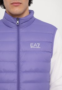 Colete roxo acolchoado com gola alta, fecho frontal e logotipo EA7 Emporio Armani. Textura suave com costura horizontal no tecido.