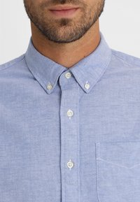 Camicia azzurro chiaro con colletto a punta larga, bottoni bianchi e tasca sul petto. Il tessuto ha una trama sottile.