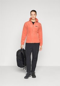 Veste en polaire corail, design à zip intégral, avec un col montant. Associée à un pantalon noir et des bottes. Individu tenant un sac à dos noir.