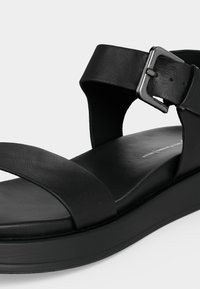 Μαύρο δερμάτινο σανδάλι με παχύ πάτο, φαρδιά λωρίδα μπροστά και λουράκι στο αστράγαλο που ασφαλίζει με ασημένια μεταλλική αγκράφα.