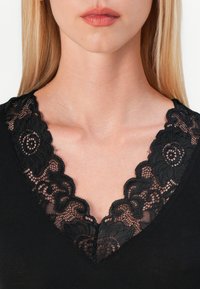 Black top featuring a V-neckline with intricate black lace trim. The lace has floral motifs and scalloped edges, contrasting with the solid fabric.