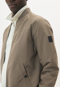 Jachetă bomber ușoară, bej, cu fermoar frontal, guler ribbed, buzunare laterale și un patch negru cu logo pe mâneca stângă.
