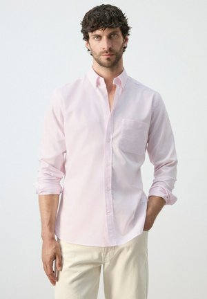 Uomo con capelli ricci scuri che indossa una camicia rosa chiaro con bottoni e maniche arrotolate e pantaloni color crema su uno sfondo semplice.