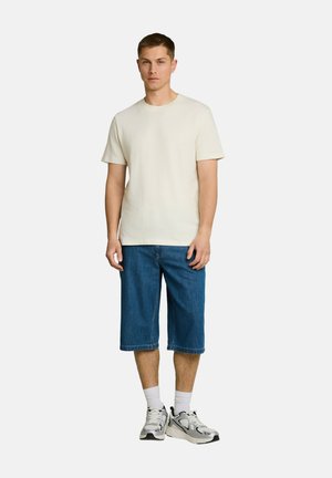 Junger Mann steht da und trägt ein schlichtes, off-weißes kurzärmeliges Hemd, kniehohe blaue Jeansshorts, weiße Socken und graue Turnschuhe vor weißem Hintergrund.