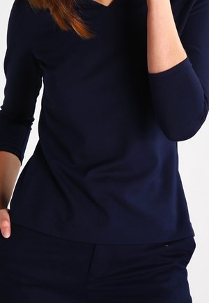 Vrouw die een marineblauw shirt met lange mouwen en marineblauwe broek draagt, met één hand in de zak en de ander dicht bij haar gezicht.