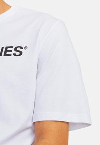 Weißes Baumwoll-T-Shirt mit kurzen Ärmeln, rundem Halsausschnitt und auffälligem schwarzen Text auf der Vorderseite. Angenehme Textur und lässige Passform.