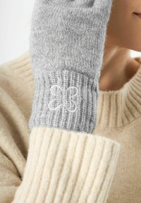 Mitten en tricot gris avec un poignet en côtes et un motif de papillon brodé blanc, porté avec un pull de couleur crème. Texture douce visible.