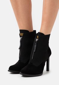 Botines negros con una combinación de ante suave y tejido de punto texturizado. Tacón alto delgado y cremalleras laterales. Logo de G-Star visible.