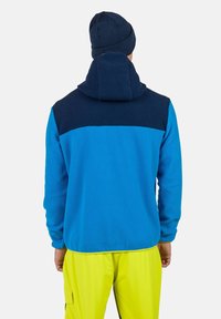 Sweater fleece sininen, tummansinisillä olkapaneeleilla. Varustettu hupulla ja tekstuurilla. Yhdistetty kirkkaan keltaisiin housuihin.