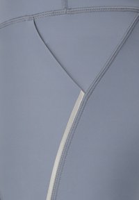 Nahaufnahme eines hellgrauen Stoffes mit detaillierter Nahtführung und einer weißen Naht, die sich kreuzende gebogene und gerade Linien zeigt.