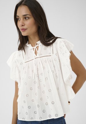 Kobieta z długimi ciemnymi włosami, ubrana w białą bluzkę z ażurowym wzorem, z falbanianymi rękawami i wiązaniem przy dekolcie, w połączeniu z niebieskimi dżinsami, na szarym tle.