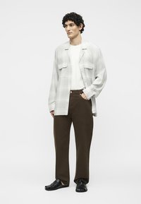 Jeune homme debout portant un t-shirt blanc, une chemise ouverte gris clair, un pantalon marron foncé et des chaussures noires, main dans la poche, fond neutre.