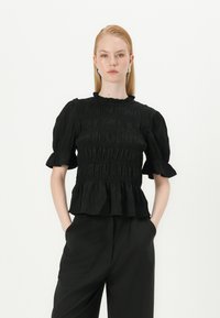 Svart smockbluse med korte pufferm, fløyelskrage og peplumkant. Stoffet har en teksturert overflate, kombinert med svarte høyt midje bukser.