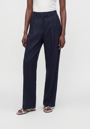 Femme portant un pantalon large taille haute bleu marine foncé, un haut blanc sans manches et des sandales plates blanches à brides sur un fond uni.
