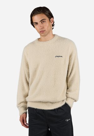 Beige fluffy sweater with a round neckline and long sleeves, featuring a small black logo on the chest. Soft texture, relaxed fit.