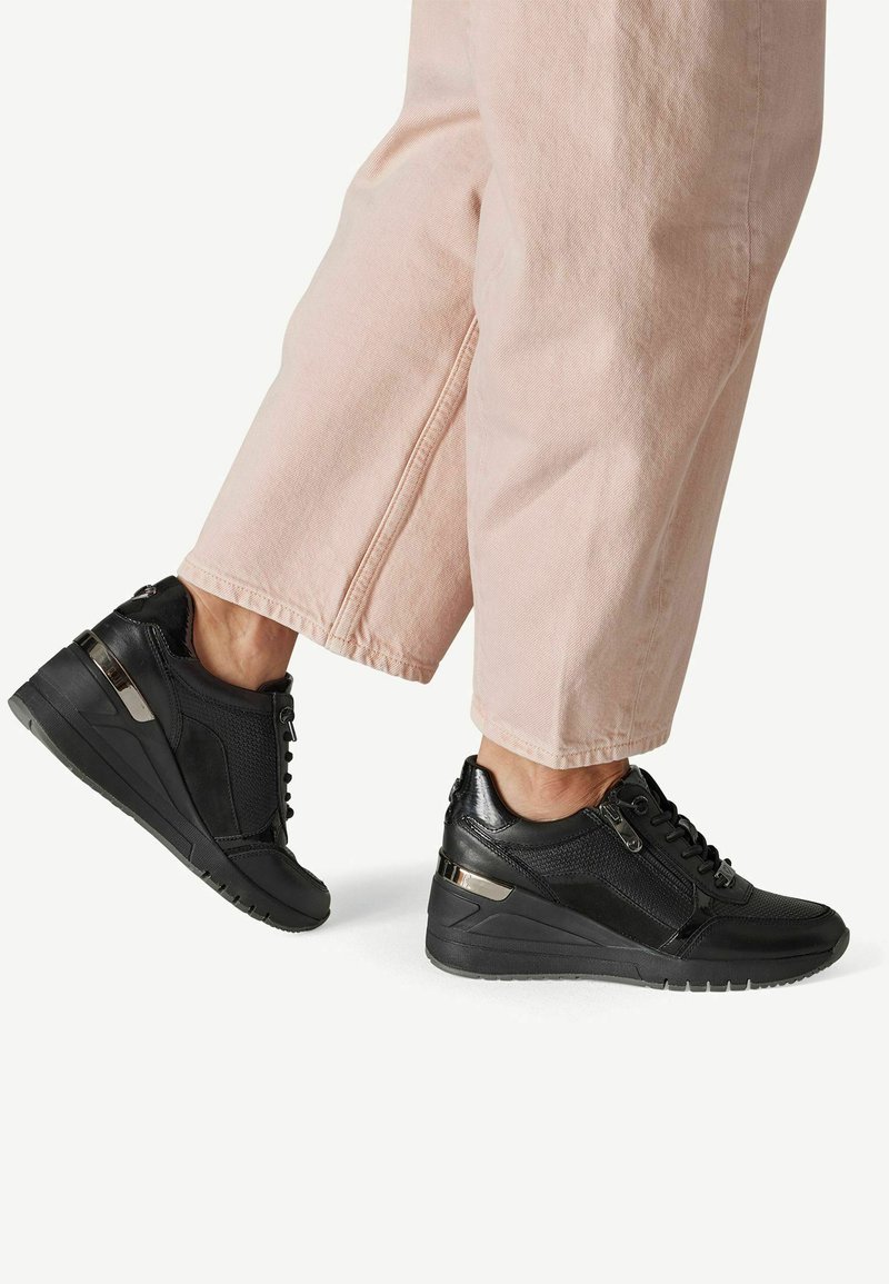Personne portant des baskets compensées noires avec une fermeture éclair latérale et des lacets, associées à un pantalon large rose clair.