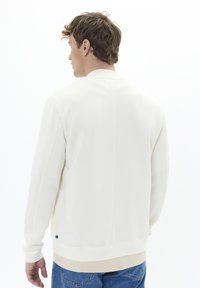 Sudadera blanca con cuello redondo, textura suave y puños ajustados. Detalle de costura notable en la parte posterior y aspecto en capas sobre vaqueros azules.
