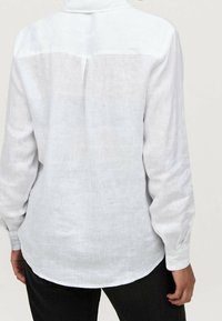 Camicia di lino bianca con maniche lunghe, cucitura a yoke sul retro e orlo curvo, con texture visibile e polsini abbottonati.