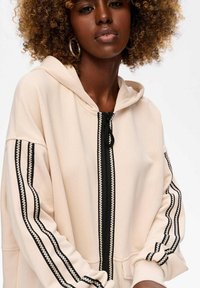 Vrouw met krullend haar draagt een beige ritsjack met zwart-witte gestreepte details op de mouwen en rits, staande tegen een effen achtergrond.