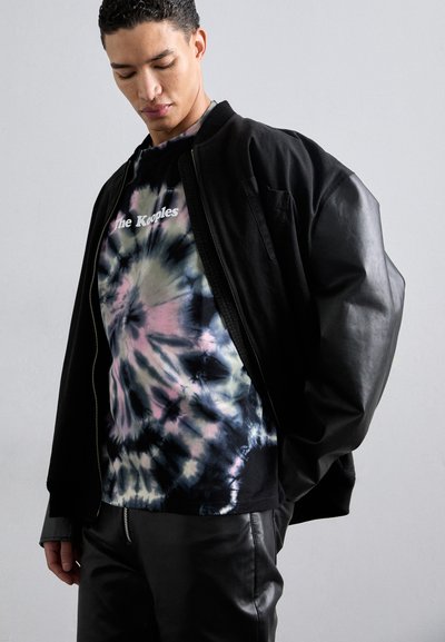 Svart bomberjakke med overdimensjonerte ermer over en tie-dye t-skjorte med pastellfarger og "The Kooples"-logo. Lærbukser synlig.