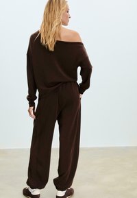 Žena s blond vlasmi, ktorá má na sebe voľný, top s odhalenými ramenami v tmavo hnedých farbách a zodpovedajúce široké nohavice, stojí na svetlej prázdnej podlahe.