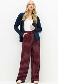 Kobieta z długimi blond włosami, ubrana w granatowy kardigan, biały top i wysoko taliowane bordowe spodnie o szerokich nogawkach z białymi paskami, stoi z jedną ręką w kieszeni.