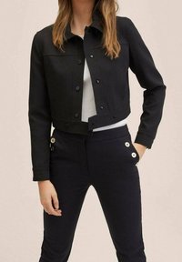 Zwarte cropped jacket van stevige stof met drukknoopjes, zijzakken en een strakke halslijn, gelaagd over een lichte top en aansluitende broek.