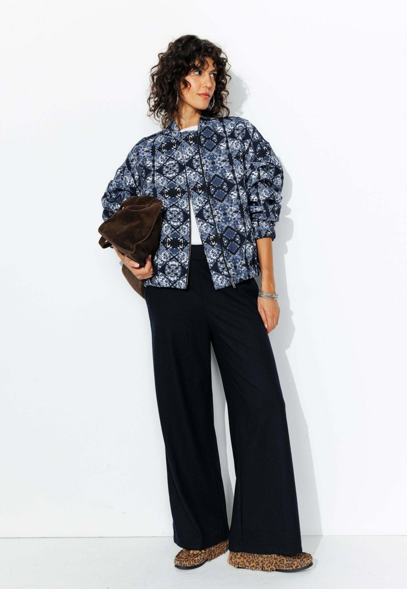 Veste bomber à motif bleu avec fermeture éclair à l'avant, associée à un pantalon large noir et des chaussures à imprimé léopard. Sac marron sous le bras.