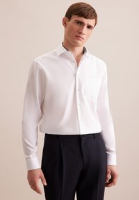 Camicia bianca a maniche lunghe con colletto e taschino piccolo sul petto, caratterizzata da un dettaglio a contrasto testurizzato sul colletto. Indossata con pantaloni scuri.