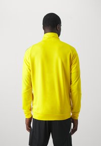 Pulover sportiv galben cu guler înalt, mâneci lungi și textură netedă, purtat cu pantaloni scurți negri. Fără logo-uri sau modele vizibile.