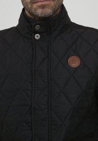 Fekete steppelt dzseki magas gallérral, fém patentos záródással és barna bőr logóval a mellrészen.