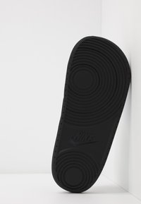 Juodas guminis sportbačių padas su apvaliais sukibimo raštais ir įspaustu Nike logotipu, pateiktas kampu prieš baltą foną.