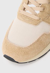 Beżowy but sportowy z teksturowaną cholewką z zamszu i siateczkowym panelem. Posiada płaską czarną podeszwę i jasnobrązowe sznurowadła.