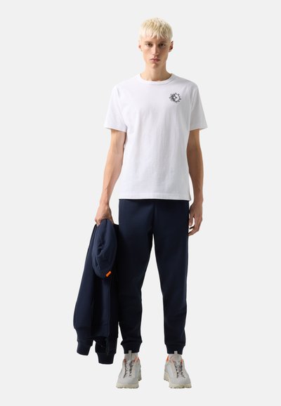 Mladý muž s krátkými blond vlasy, který má na sobě bílé tričko s grafikou, tmavě modré tepláky a bílé tenisky, v jedné ruce drží tmavě modrou bundu.