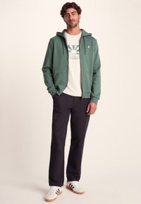 Green zip-up hoodie with a drawstring and front pockets, layered over a cream graphic t-shirt, paired with black pants and white sneakers.